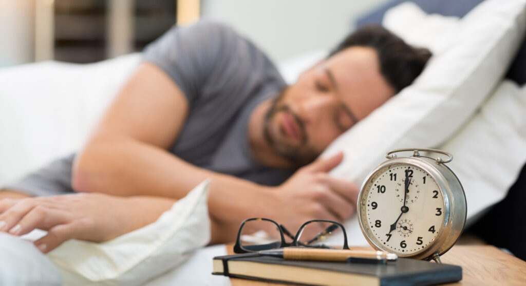 Man asleep in bed with alarm clock and glasses on nightstand